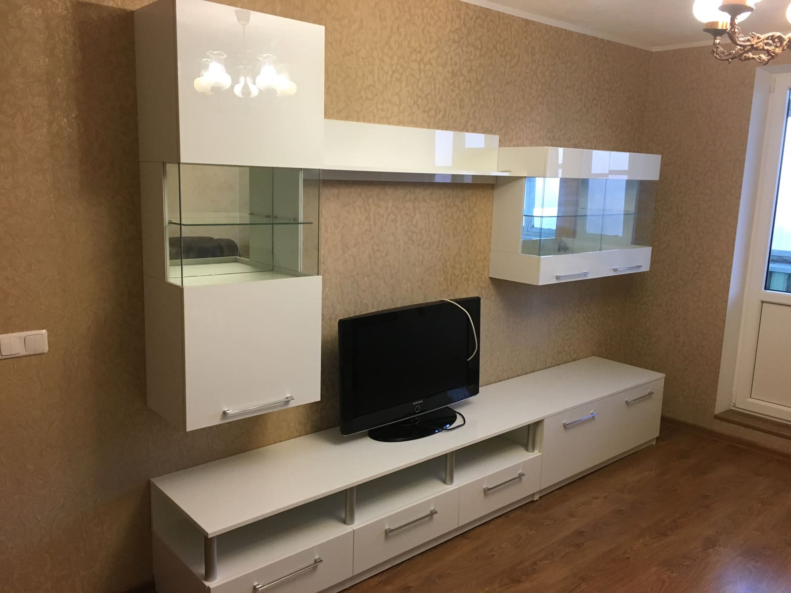 Svetainės baldų komplektas: baltos blizgios spintelės po televizoriumi ir pakabinama vitrina su apšvietimu